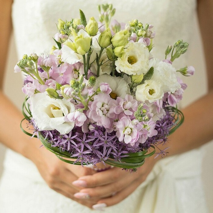 In a close-up evocative of a Deptford wedding morning, this bouquet radiates fresh artistry and gentle vibrancy. The eye is drawn first to elegant white Lisianthus, their soft, undulating petals brushed with the faintest touch of green at the centre, paired with pale pink and lilac stock blossoms whose multi-petaled forms suggest cascading sweetness. Among these, tight green buds dot the arrangement, signifying hope and anticipation, while the base is dramatically encircled by a band of rich purple Allium-spiky star-like forms that inject both structure and contemporary flair. Encasing the composition, long, looping strips of glossy bear grass weave thoughtfully through the arrangement, underlining both its freshness and cohesiveness. The bouquet rests gently in the bride's hands-delicately manicured and poised-while a soft veil of white lace from the dress provides an ethereal backdrop, blurred just out of focus, reminiscent of a Deptford wedding along Evelyn Street. This lush arrangement offers a harmonious balance of old and new, echoing Deptford's renewal and artistic energy, making it ideal for brides seeking a modern yet timeless floral statement.